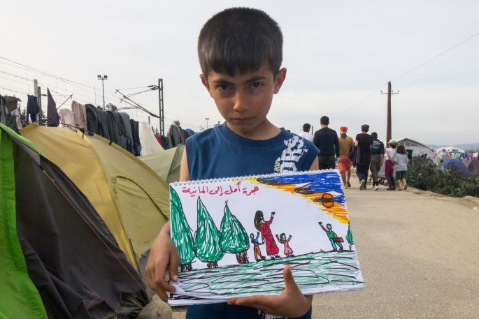 An unnamed Syrian refugee boy shows his painting book where he has painted scenes from the war at Syria and his travel to Idomeni refugee camp on the Greek-Macedonia borders. The writing says "hope in Germany" and the painting shows the father leaving and his family waving goodbye.  All photographs: Kostas Pikoulas


