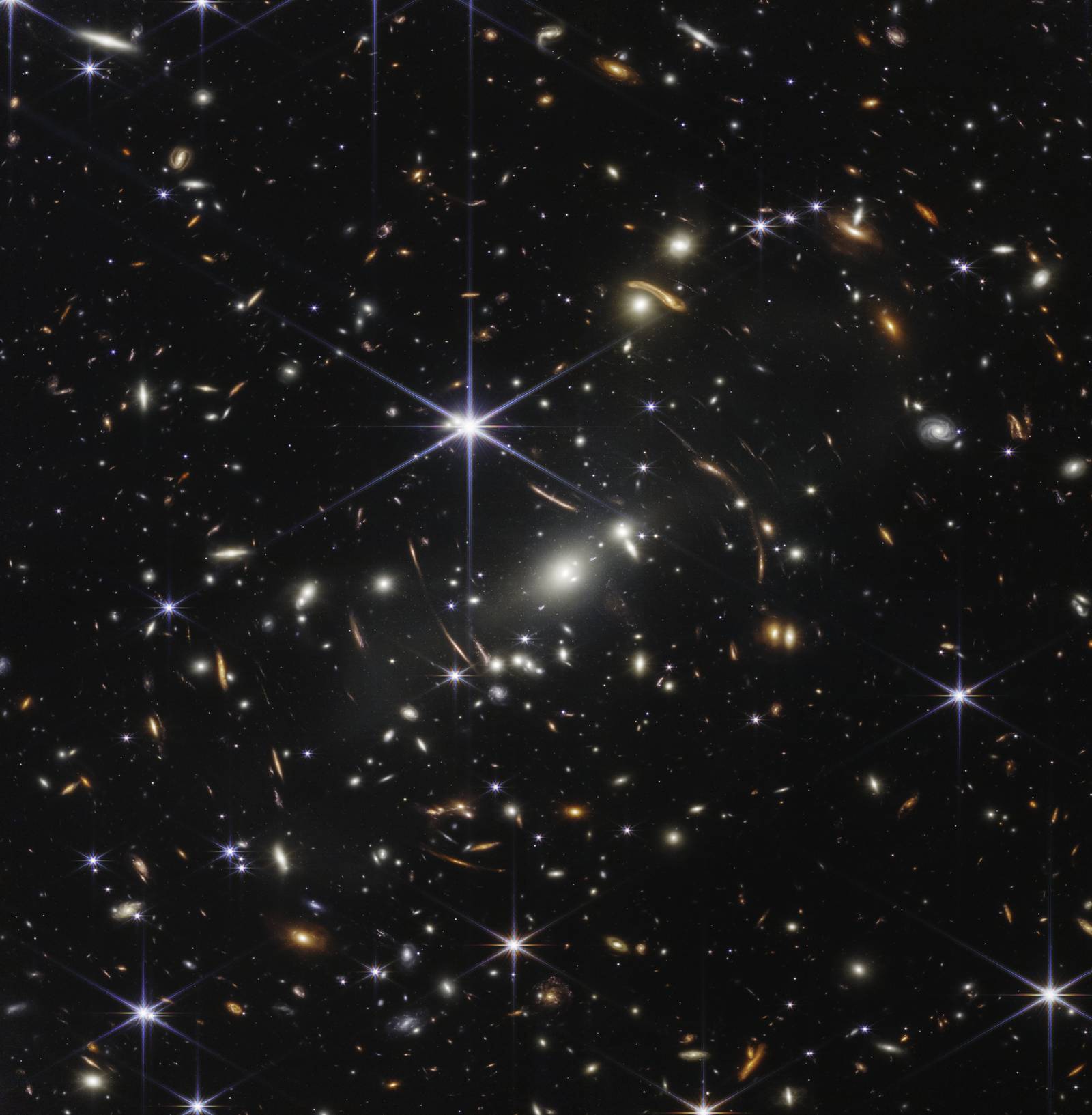 Thousands of small galaxies appear across the James Webb Space Telescope’s first deep field image. Their colours vary. Some are shades of orange, while others are white. Most appear as fuzzy ovals, but a few have distinct spiral arms. In front of the galaxies are several foreground stars. Most appear blue, and the bright stars have diffraction spikes, forming an eight-pointed star shape. There are also many thin, long, orange arcs that curve around the centre of the image.