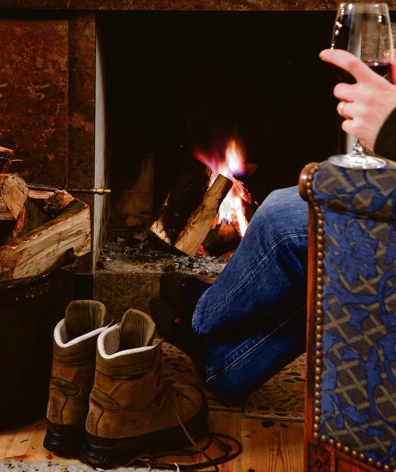 Man relaxing with a glass of red wine by the fireplace after a long hike; his boots off, next to him, warming up