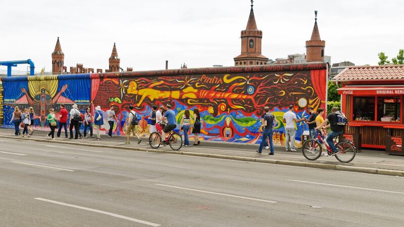 The Berlin Wall at the Muehlenstrasse. The 3m-high wall separated the city Berlin from 1961 - 1989. Today the remains serve as a memorial and offer artists a space. East Side Gallery the longest wall gallery. The remains of the wall are very popular with tourists. Young people on front of the wall. In the background to see the towers of the Oberbaumbruecke.”
